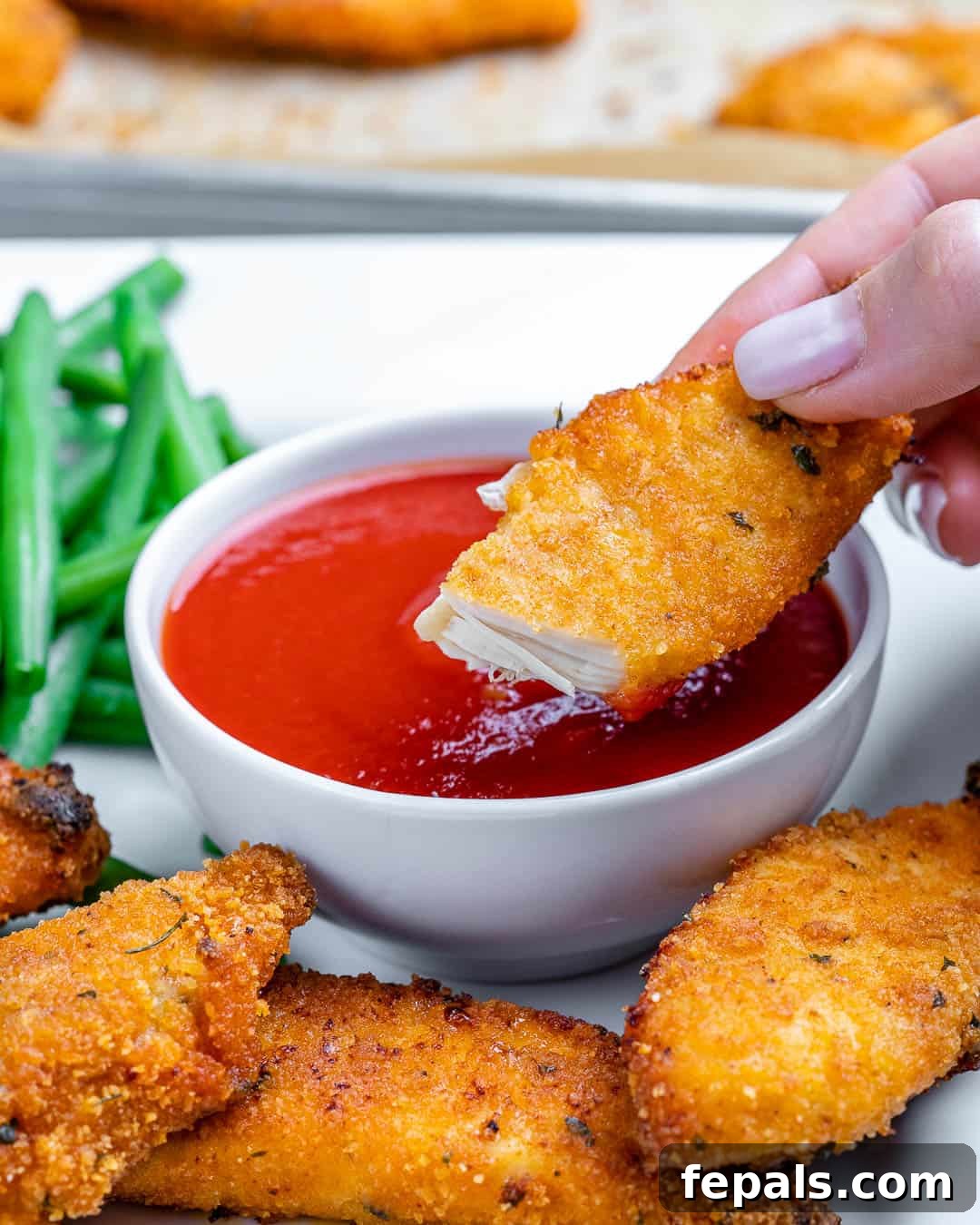 A hand dipping a crispy oven-fried chicken tender into a bowl of vibrant red ketchup.