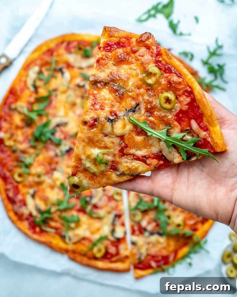 A hand holds a single, perfectly sliced piece of low-carb pizza, featuring fresh arugula and vibrant green olives, above the rest of the pizza.