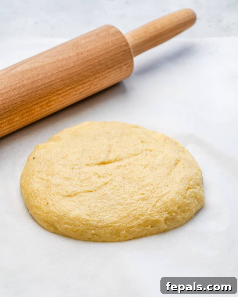 Smooth fathead low-carb pizza dough is rolled out thinly between two sheets of parchment paper, next to a rolling pin, ready for the oven.