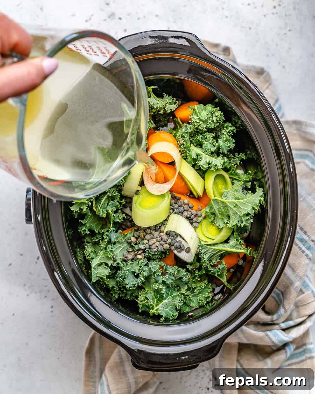 Nourishing Vegan Lentil Soup 5 Rich vegetable broth being poured into a slow cooker already filled with vibrant kale and other prepped vegetables, symbolizing the start of a wholesome lentil soup.