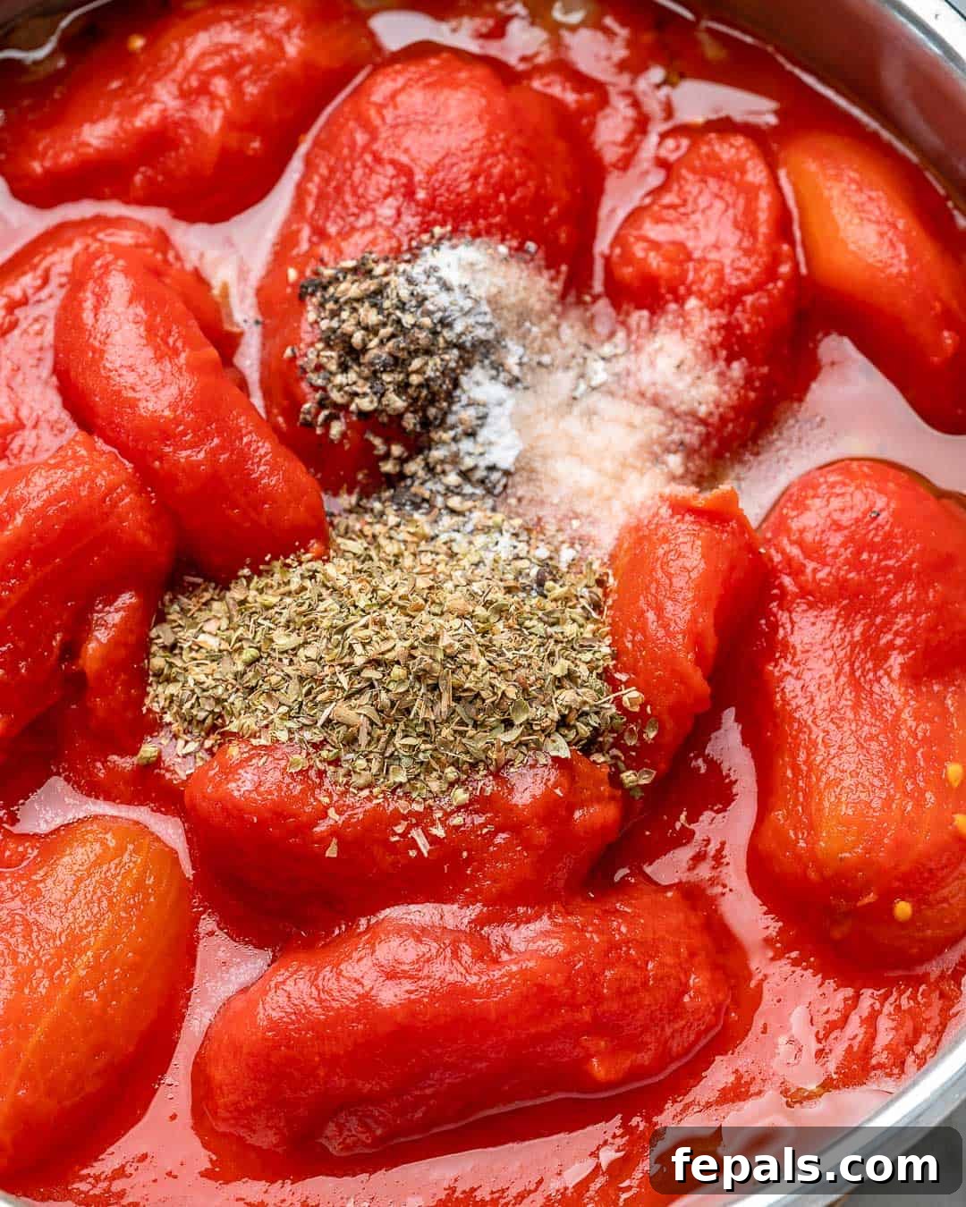 Canned whole peeled tomatoes being poured into a pan with sautéed onions and garlic, seasoned with oregano, red pepper flakes, and salt.