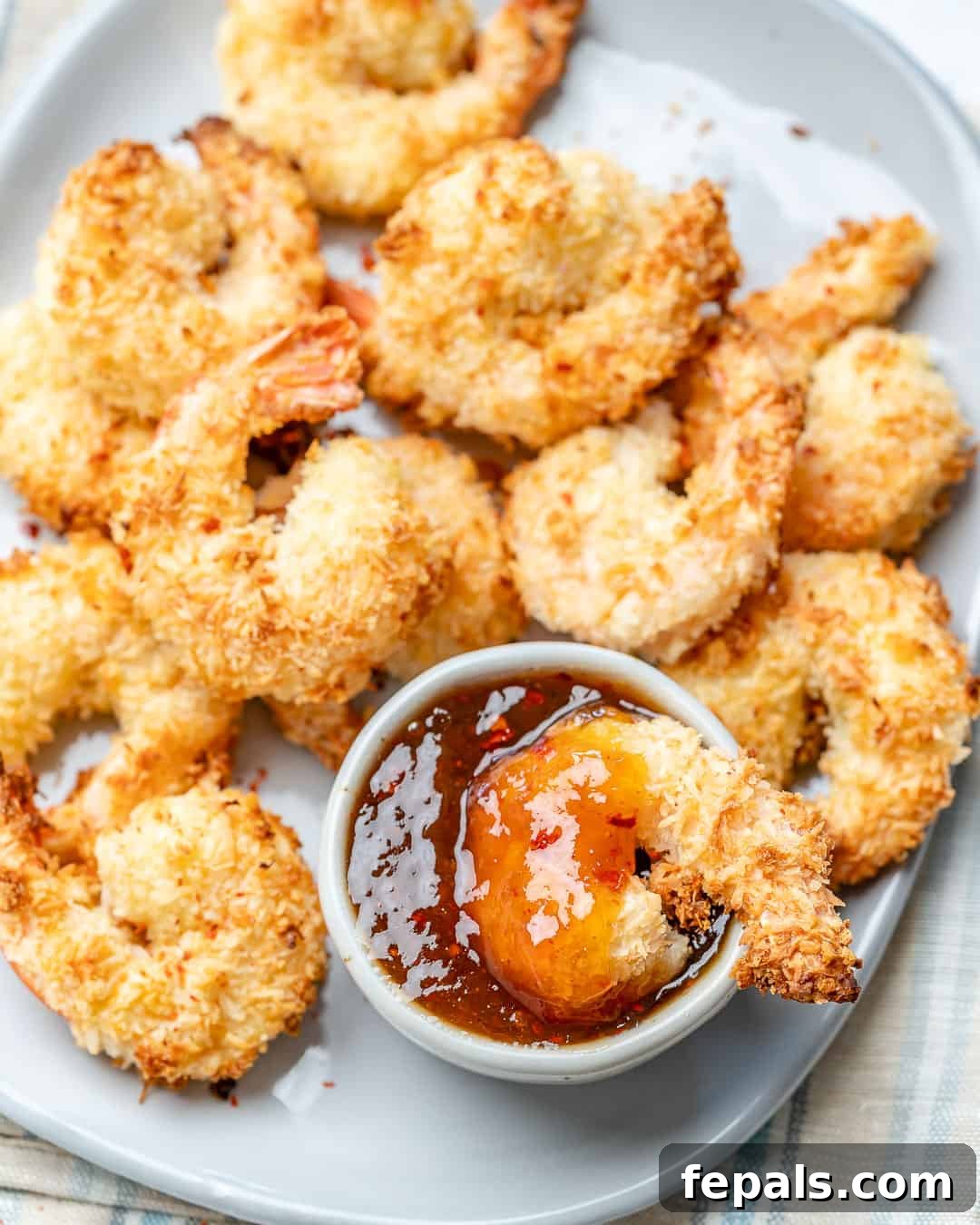 a single coconut shrimp in a dipping sauce bowl next to more baked coconut shrimp in a dish.