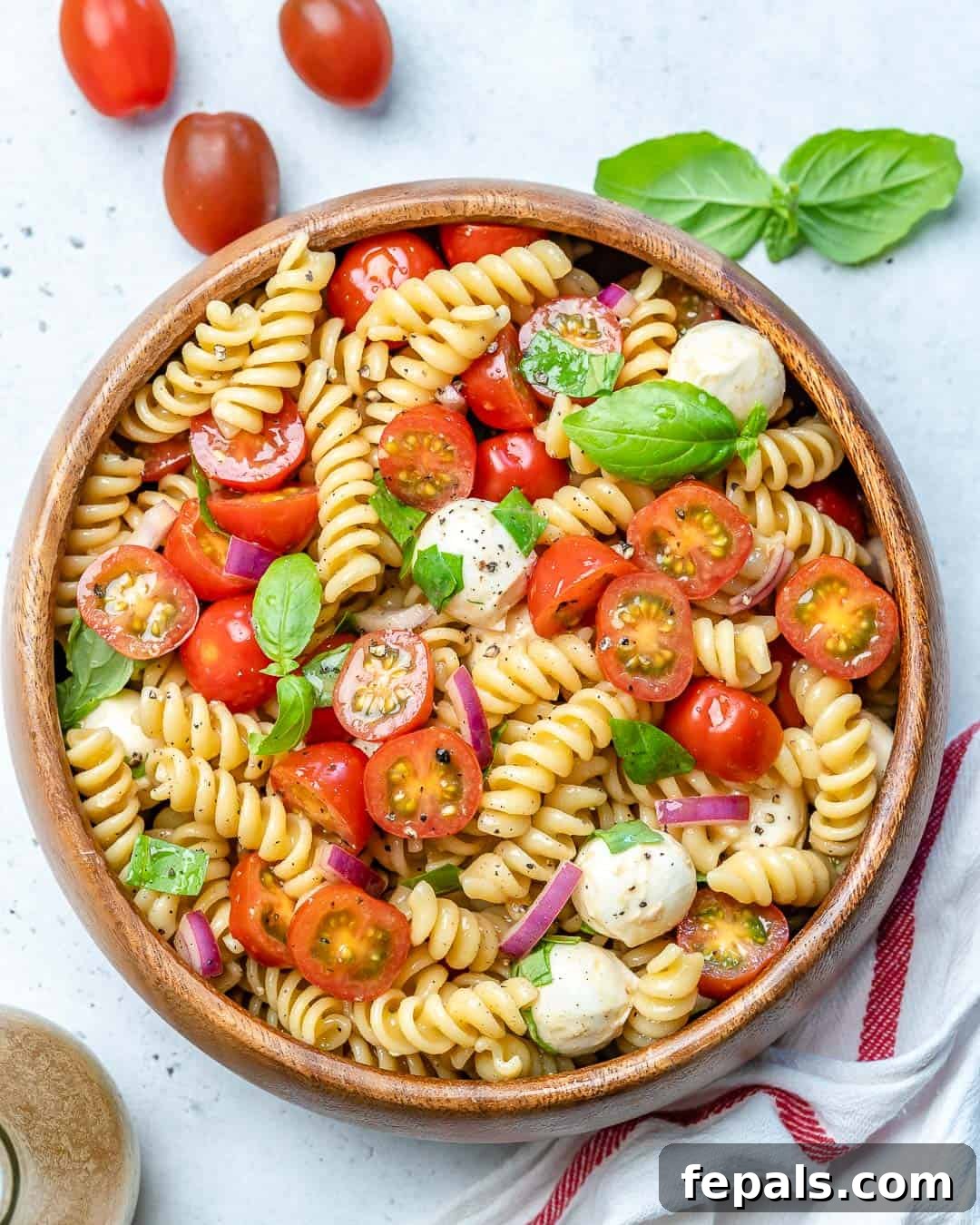 Caprese pasta salad in a round wooden bowl, garnished with fresh basil leaves and a drizzle of balsamic glaze.