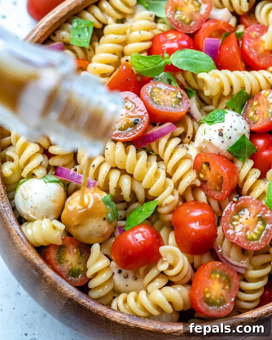 Close-up view of balsamic vinaigrette being drizzled over a freshly prepared Caprese pasta salad in a wooden bowl.