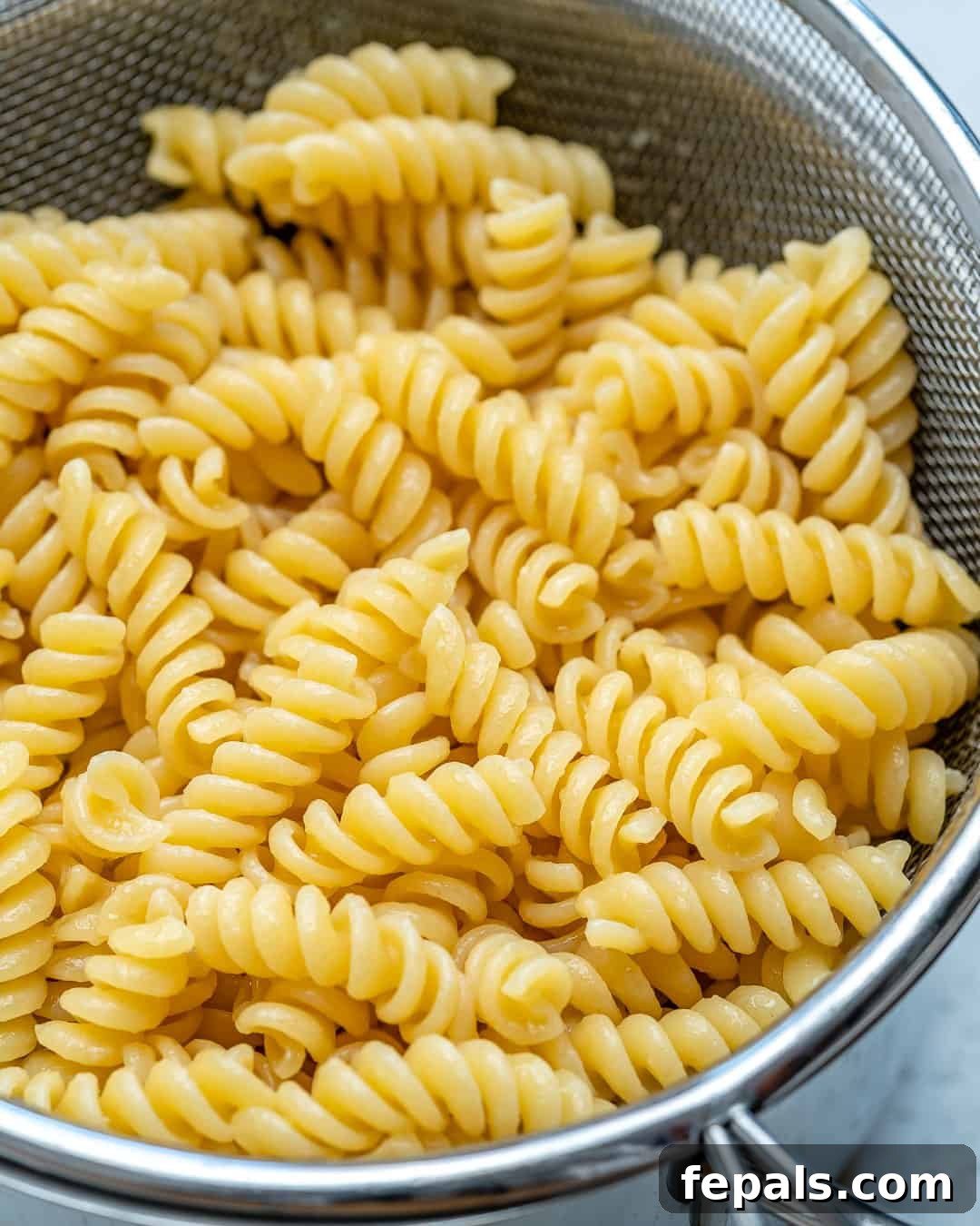 Cooked short pasta draining in a colander under cold running water, prepared for pasta salad.