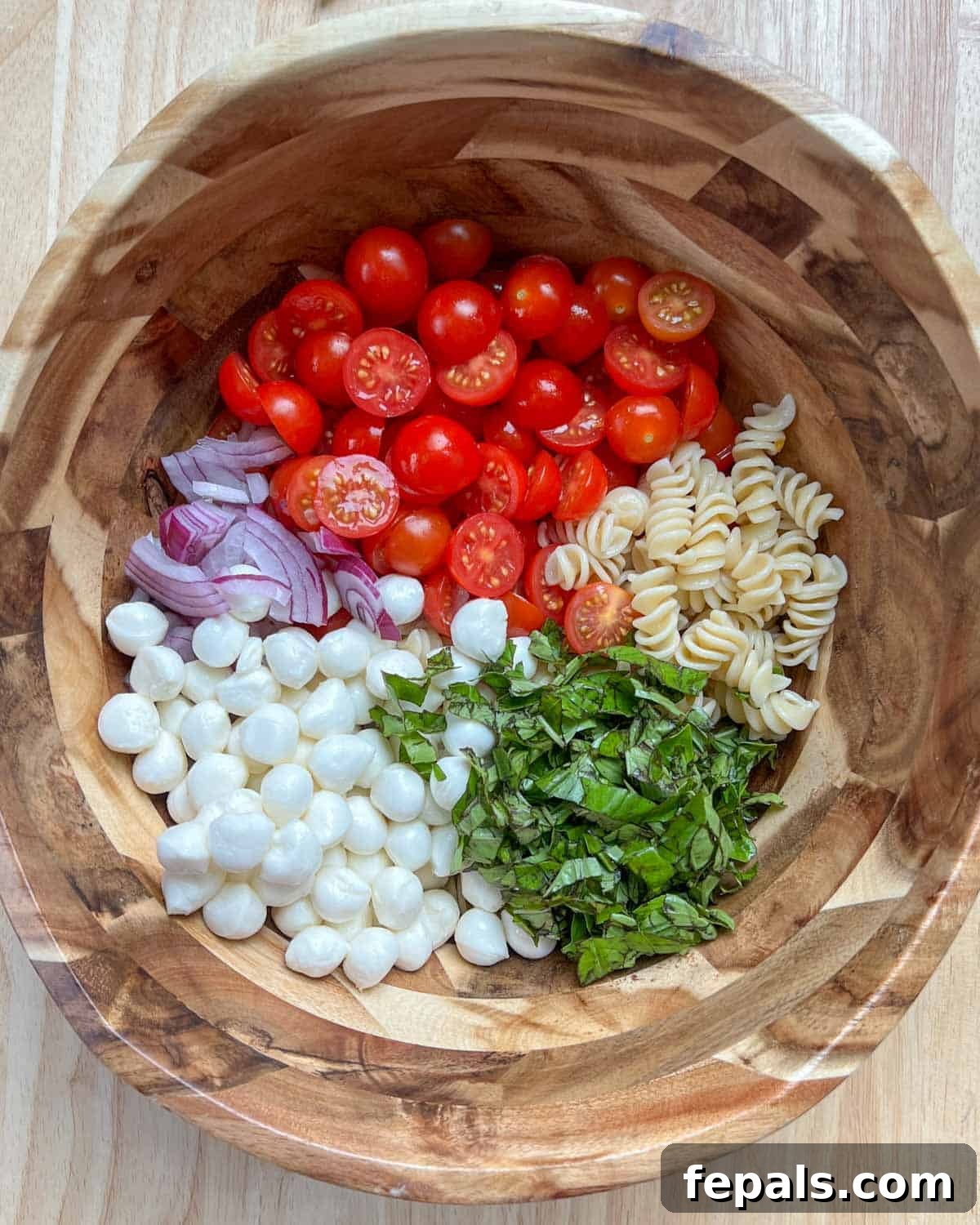 A large mixing bowl filled with cooked pasta, halved cherry tomatoes, sliced red onions, and fresh basil, ready for the dressing.