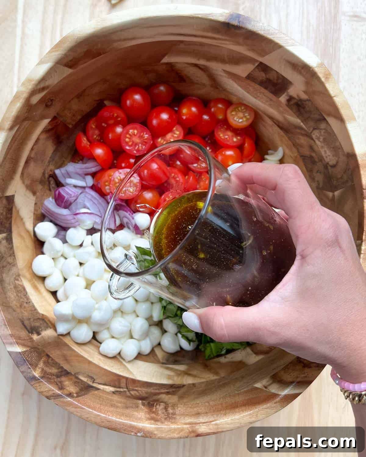 A hand pouring balsamic vinaigrette from a jar over the Caprese pasta salad ingredients in a mixing bowl.