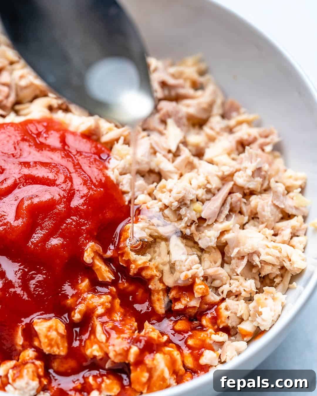 Close-up of shredded chicken being tossed with a rich, red buffalo sauce in a mixing bowl.