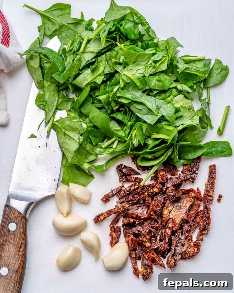 Chopped basil, finely chopped sun-dried tomatoes, and whole garlic cloves arranged neatly on a wooden cutting board, ready for the sauce.
