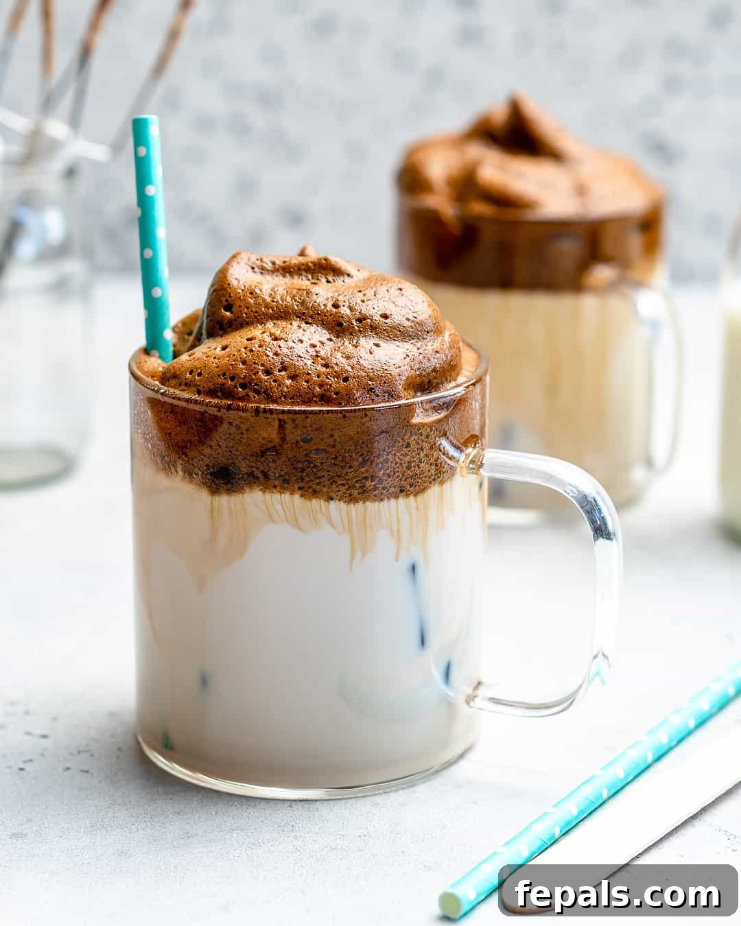 A beautifully presented whipped coffee in a transparent glass cup, adorned with a straw, showcasing its distinct layers of creamy froth and milk. The perfect homemade gourmet coffee.