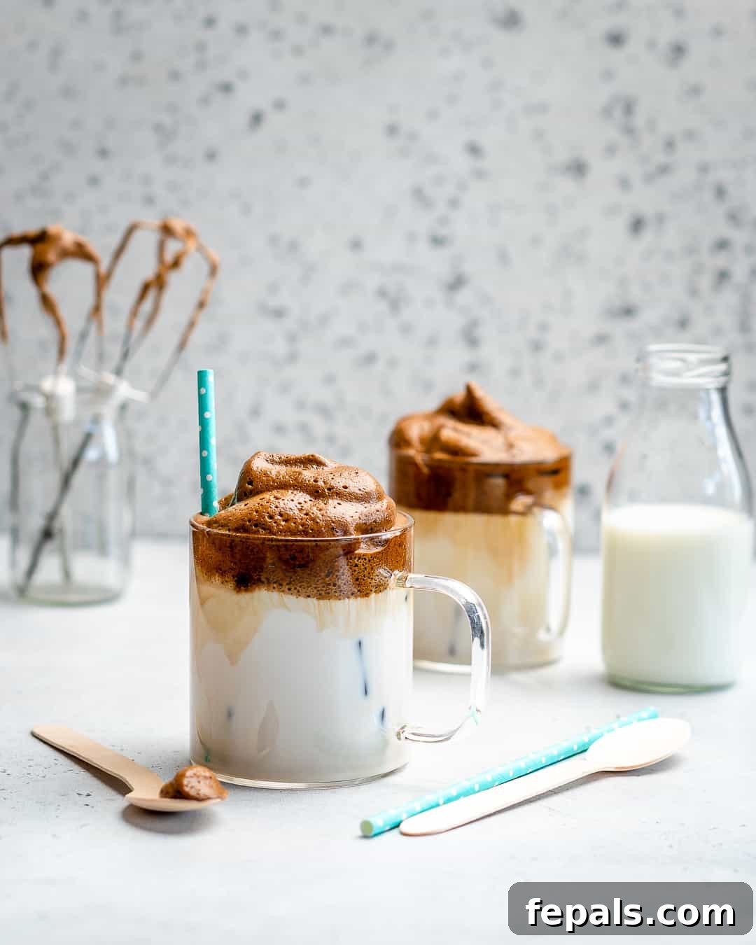 A captivating side-shot of two tall glasses of whipped coffee, one with a straw, positioned next to a glass bottle of milk. The scene suggests a refreshing, gourmet coffee experience at home.