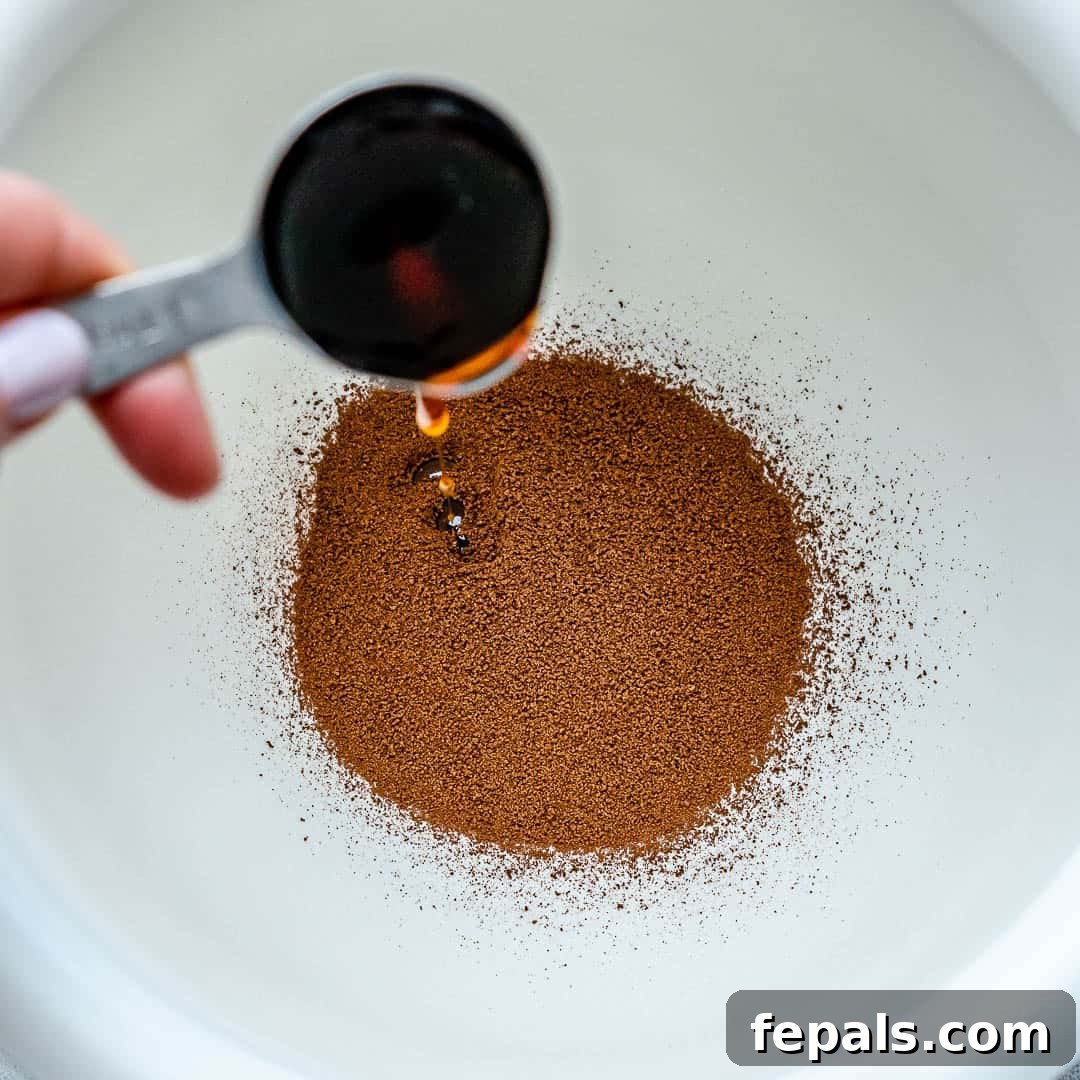 Close-up of maple syrup being poured over instant coffee and sugar in a mixing bowl, illustrating the initial step of making whipped coffee.