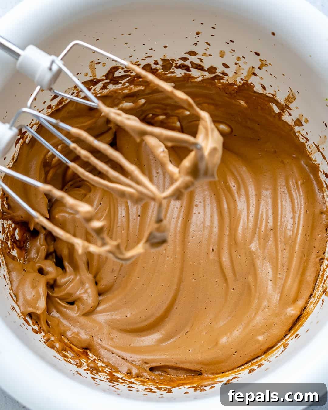 A hand mixer elevated above a bowl of perfectly whipped coffee, showcasing the thick, airy, and caramel-colored froth that has formed.