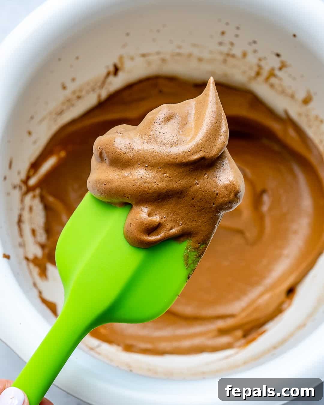 A green spatula lifting a generous portion of whipped coffee froth from a bowl, highlighting its creamy and airy texture.