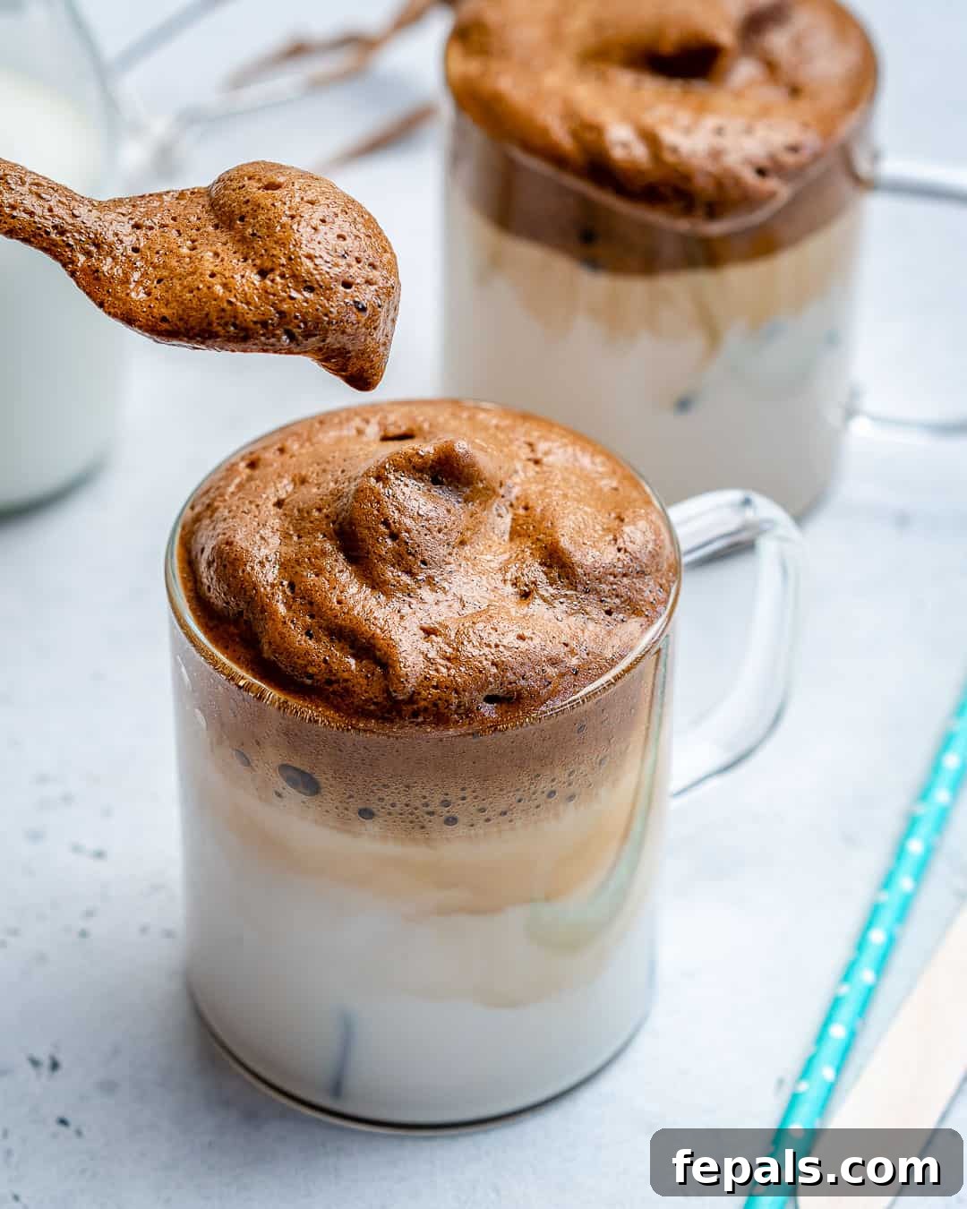 A close-up shot of a spoon holding a generous dollop of whipped coffee froth, poised next to a cup containing the layered coffee and milk drink. Emphasizes the rich, creamy texture.