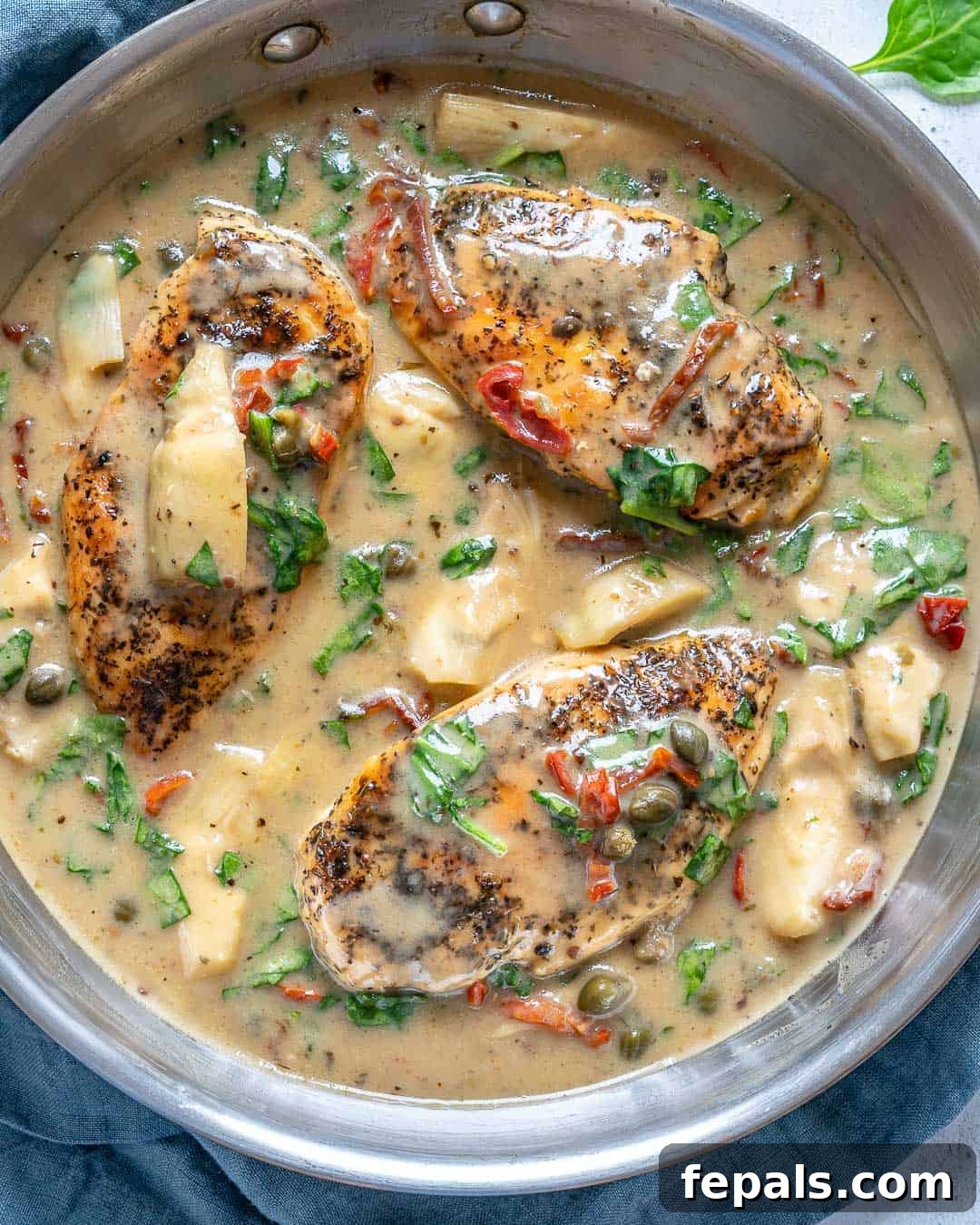 Creamy Tuscan Chicken Delight 6 Three perfectly cooked chicken breasts submerged in a rich, creamy Tuscan sauce, brimming with sun-dried tomatoes, spinach, and artichokes in a skillet.