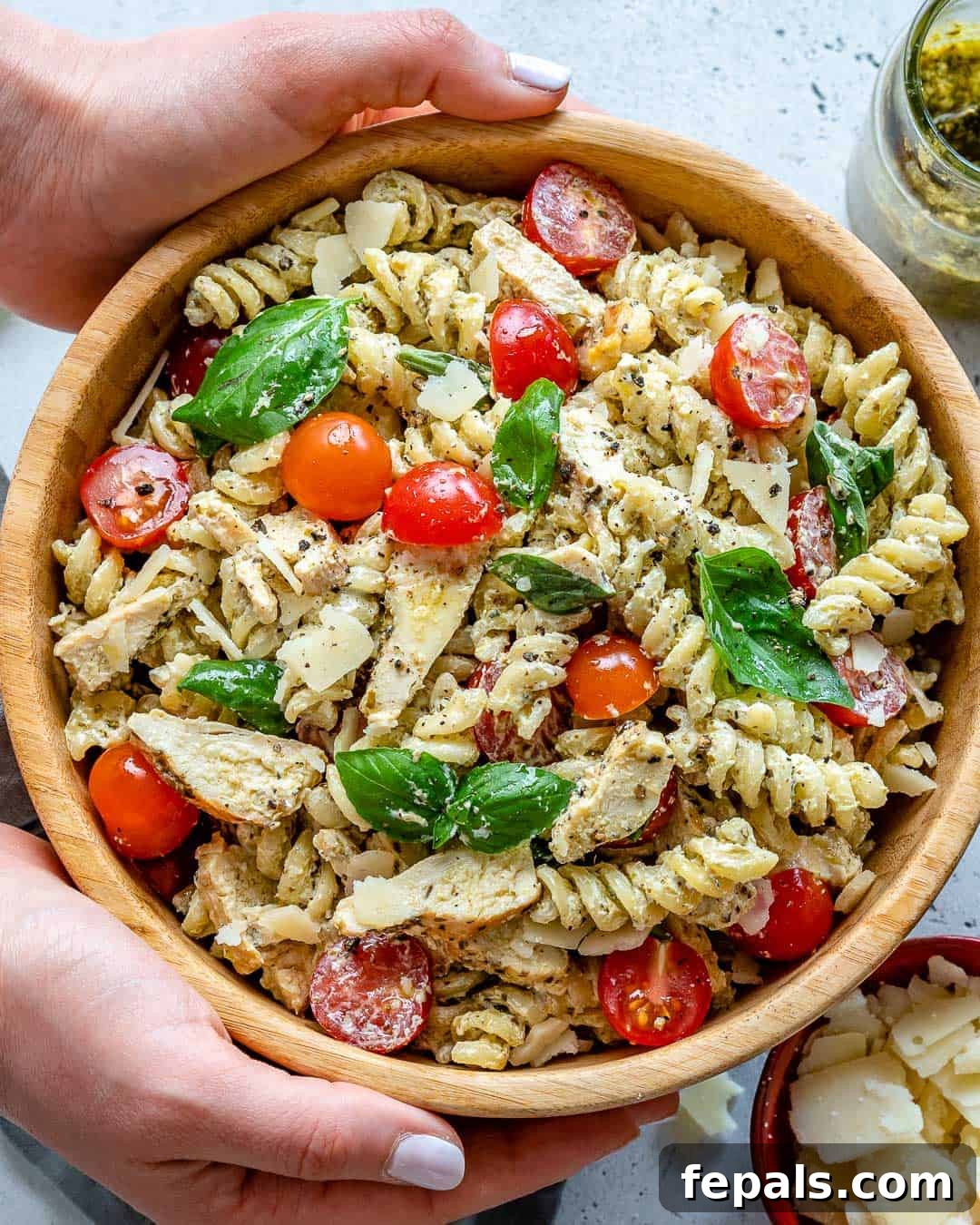 A hand holding a small white bowl filled with creamy chicken pesto pasta salad, showcasing the vibrant colors and fresh ingredients.