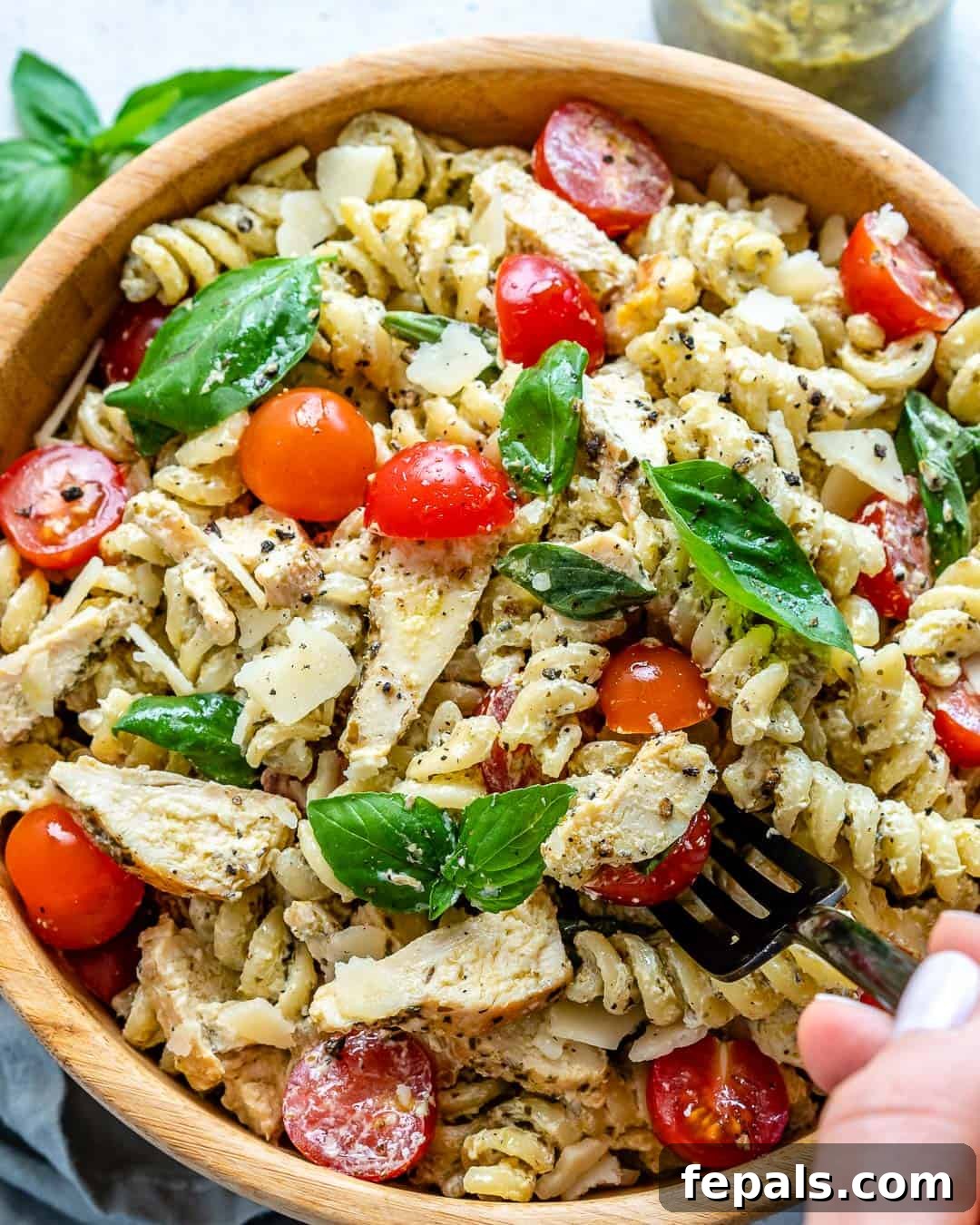 A hand reaching with a fork into a bowl of creamy chicken pesto pasta salad, perfectly showing the texture and ingredients.