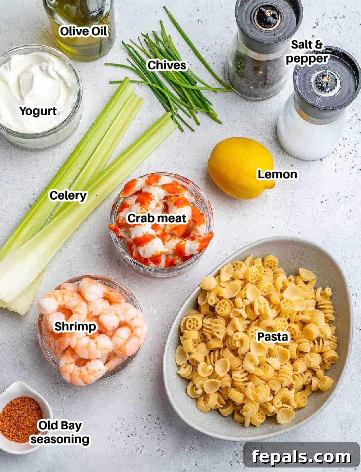 A flat lay of fresh ingredients laid out on a surface, including pasta, shrimp, crab meat, celery, chives, lemon, Greek yogurt, and Old Bay seasoning, ready to make shrimp and crab pasta salad.