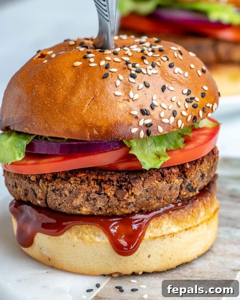 side shot of a black bean vegan burger on a surface with a knife in the middle of the burger and BBQ sauce dripping out of the burger