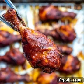 Hand holding one chicken drumstick over a pan with many baked drumsticks, illustrating the perfect golden brown color and glaze.