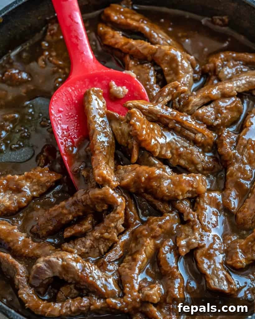 Close-up view of seared beef strips coated in a rich Mongolian sauce, cooking in a skillet with a red spatula.