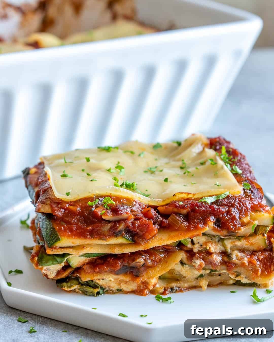 A perfectly portioned slice of golden-brown vegan zucchini lasagna, showcasing the distinct layers of pasta, creamy filling, vibrant spinach, and tender zucchini, served on a white plate.