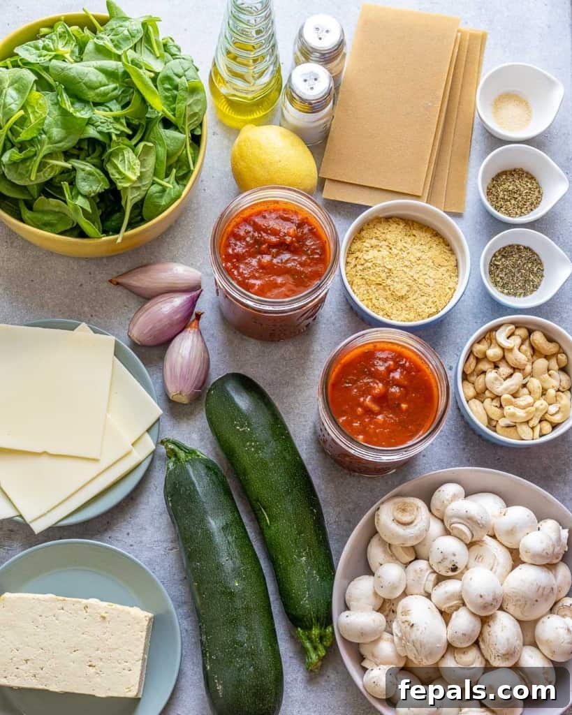 A colorful array of fresh ingredients for vegan zucchini lasagna laid out on a kitchen counter, including whole zucchini, firm tofu, button mushrooms, spinach, cashews, and marinara sauce.