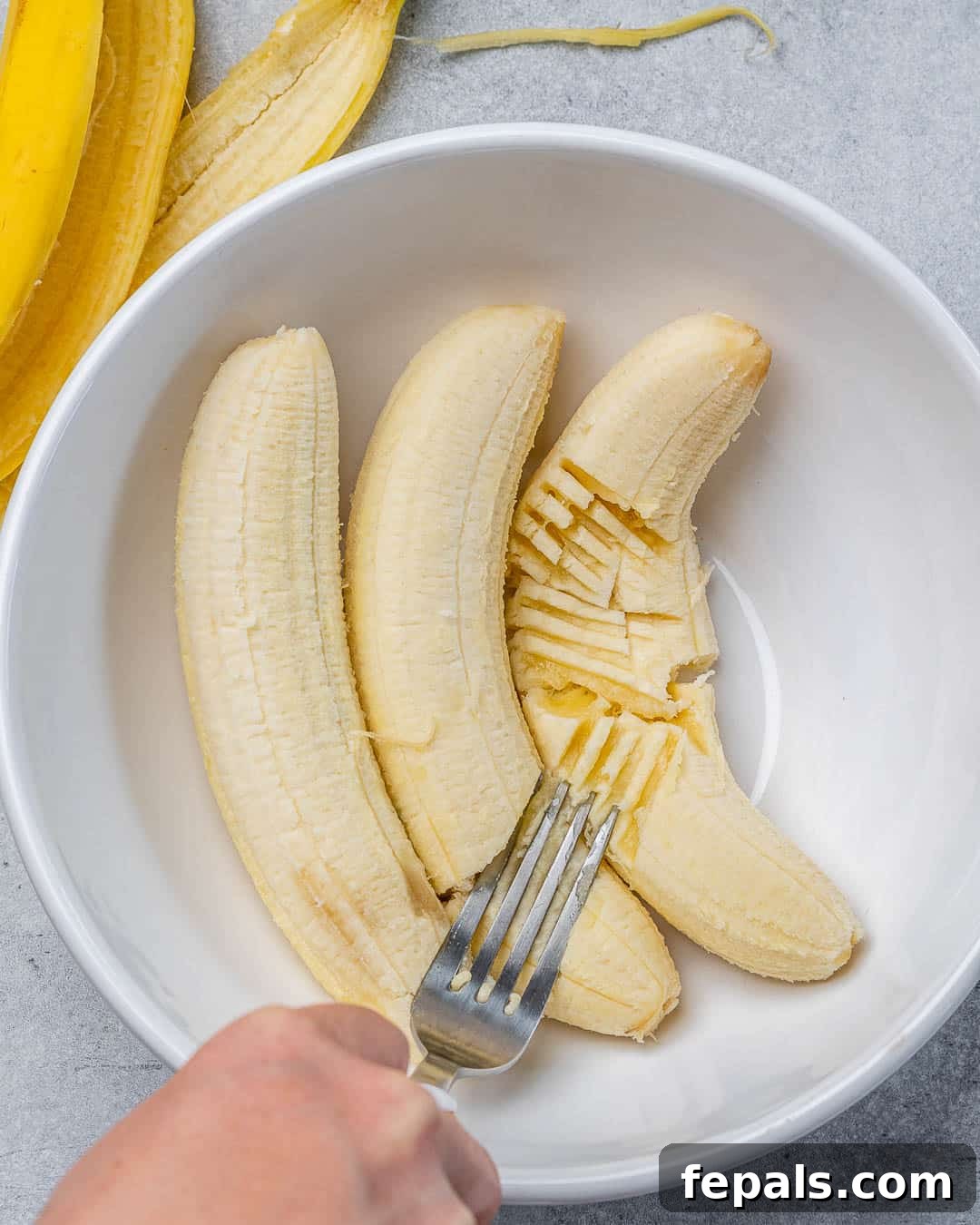 A fork mashing ripe bananas in a white bowl, showing the process of preparing the wet ingredients.