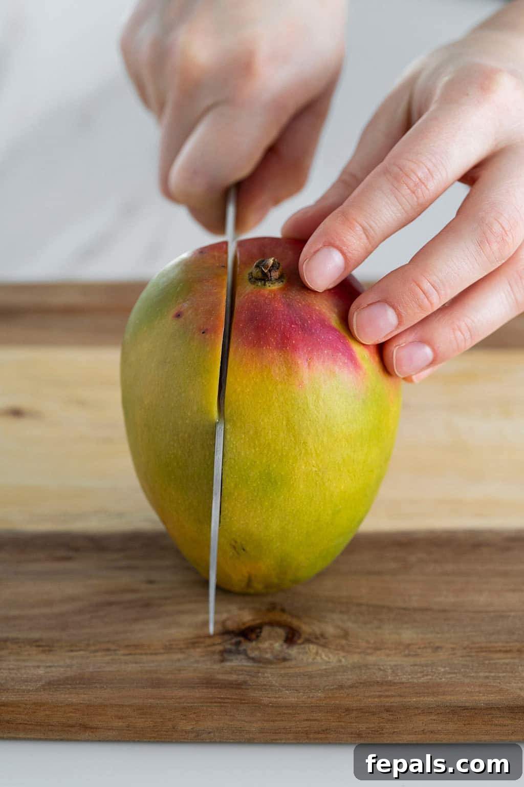 Mango with stem side up and knife slicing flesh side down, showing the initial cut