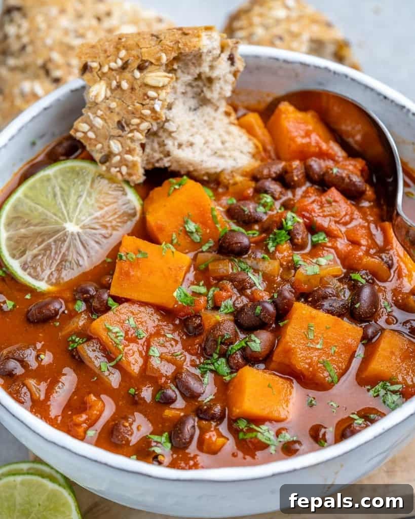 Comforting Sweet Potato Chili 7 A close-up shot of a white bowl filled with sweet potato chili, garnished with lime wedges and fresh cilantro, with a piece of crusty bread dipped in, highlighting its comforting appeal.