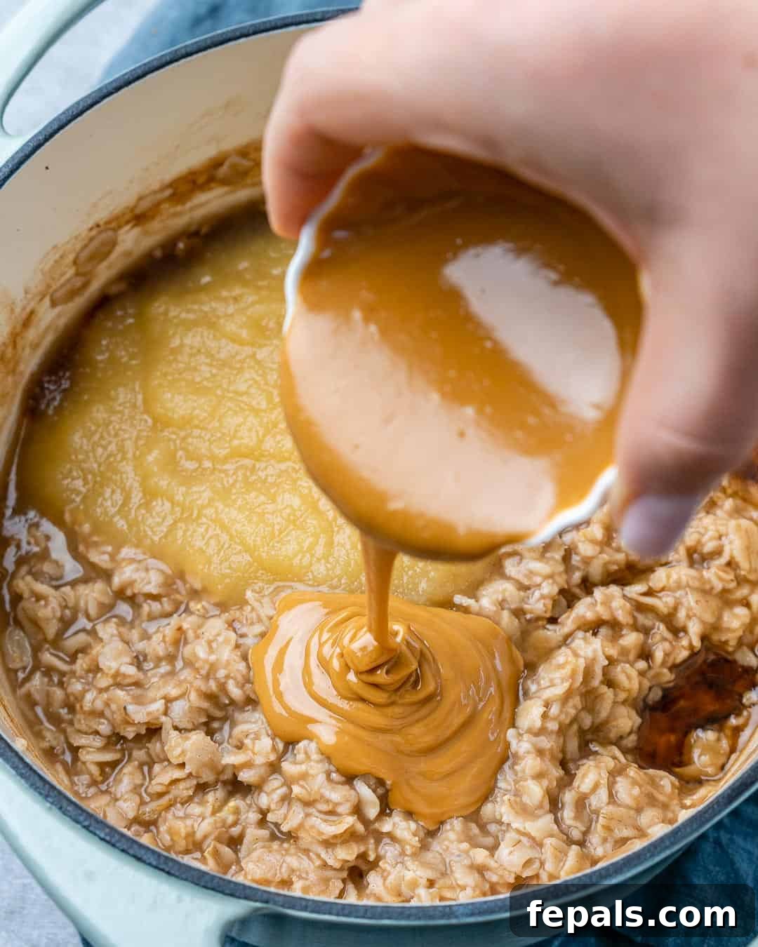 Nutty Oatmeal Morning Fuel 6 A hand pouring creamy peanut butter into a pot of cooked oats.