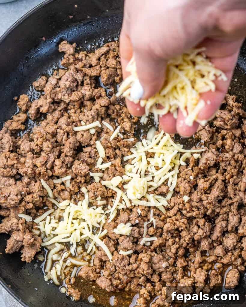 Rancher's Rustic Queso 5 Shredded cheese being carefully added to a skillet containing browned ground beef and rich beef stock, beginning the process of creating a creamy queso base