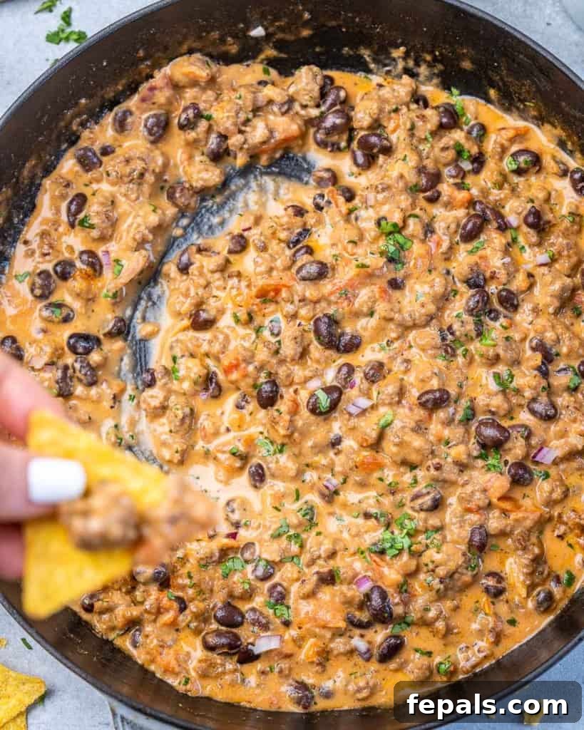 Rancher's Rustic Queso 9 A sturdy tortilla chip generously scooping up a large, satisfying portion of creamy, meaty cowboy queso dip from a rustic serving bowl, showcasing its rich texture