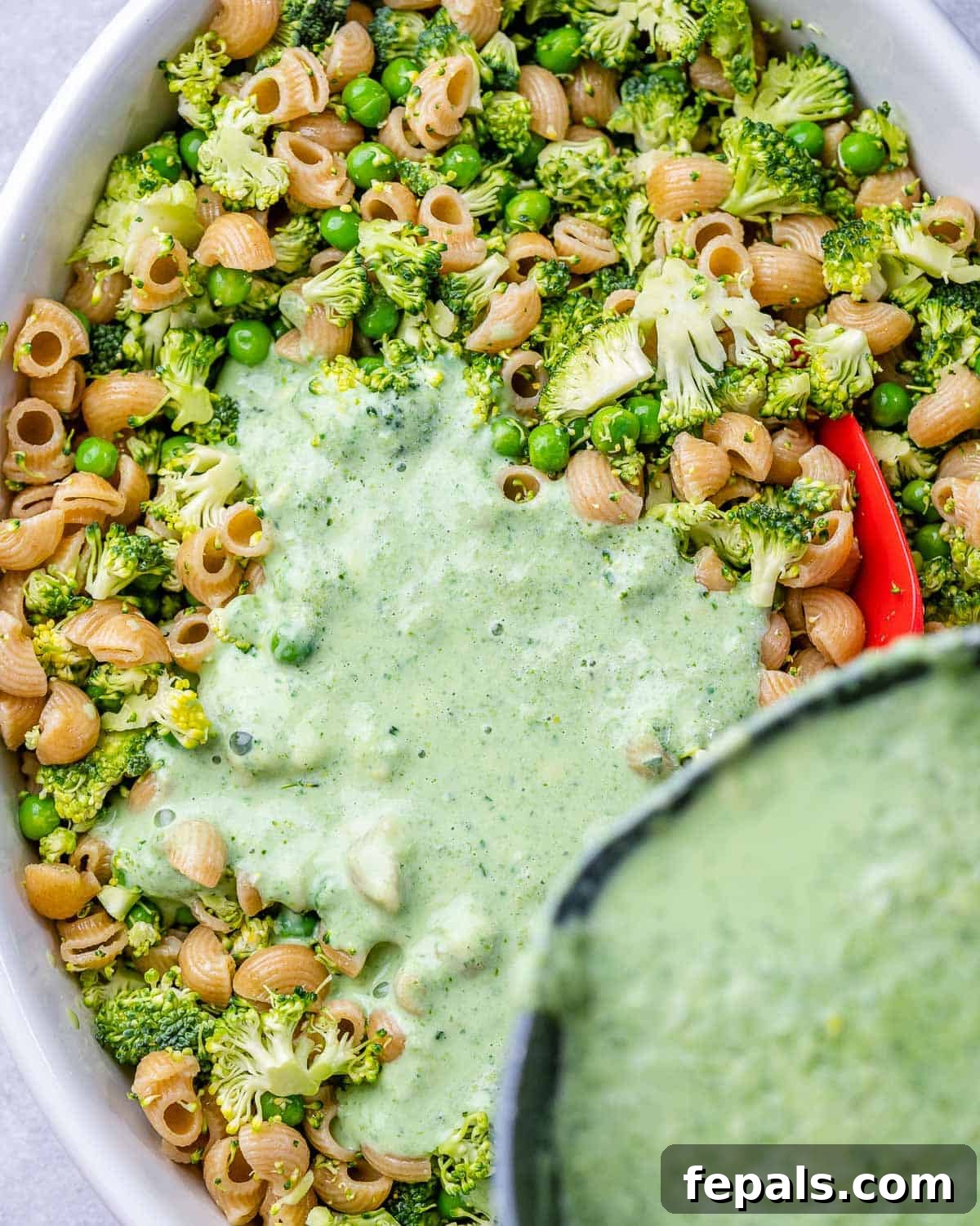 Pouring the creamy green cheese sauce over the pasta and vegetables in a baking dish.