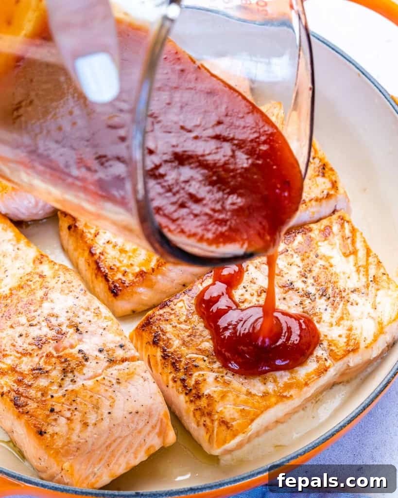Fire-Kissed Salmon 7 A hand pouring thick, rich BBQ sauce over pan-seared salmon fillets in a skillet, ensuring even coating.