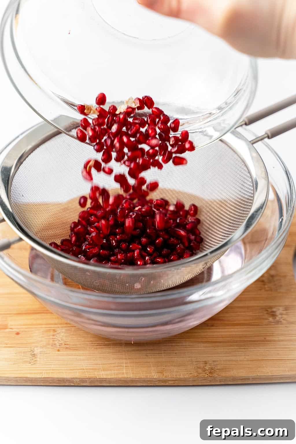 How to Cut a Pomegranate (Easy) Pomegranate arils being poured from a bowl into a mesh strainer, rinsing under running water.