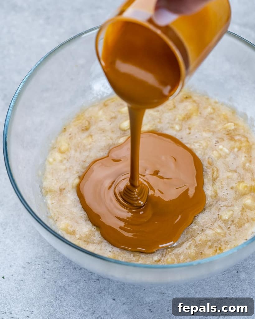 Chewy Raisin Oat Delights 5 Creamy peanut butter being poured over mashed ripe bananas in a mixing bowl, illustrating the initial steps of the recipe.