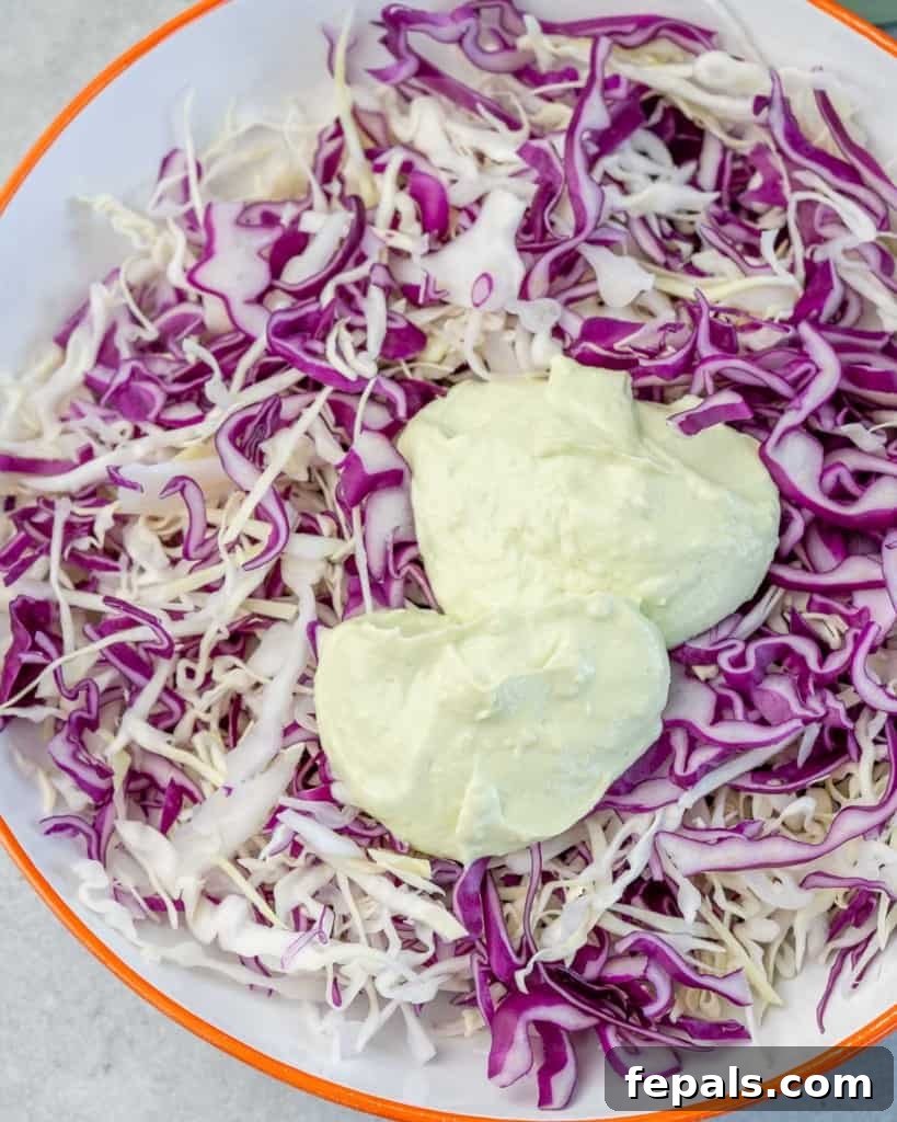 Vibrant shredded cabbage being tossed with a generous amount of creamy avocado yogurt sauce in a bowl, forming the coleslaw base.