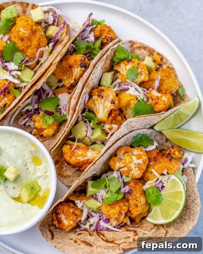 Top-down view of four assembled cauliflower tacos arranged neatly on a white plate, accompanied by a small bowl of creamy avocado dip.