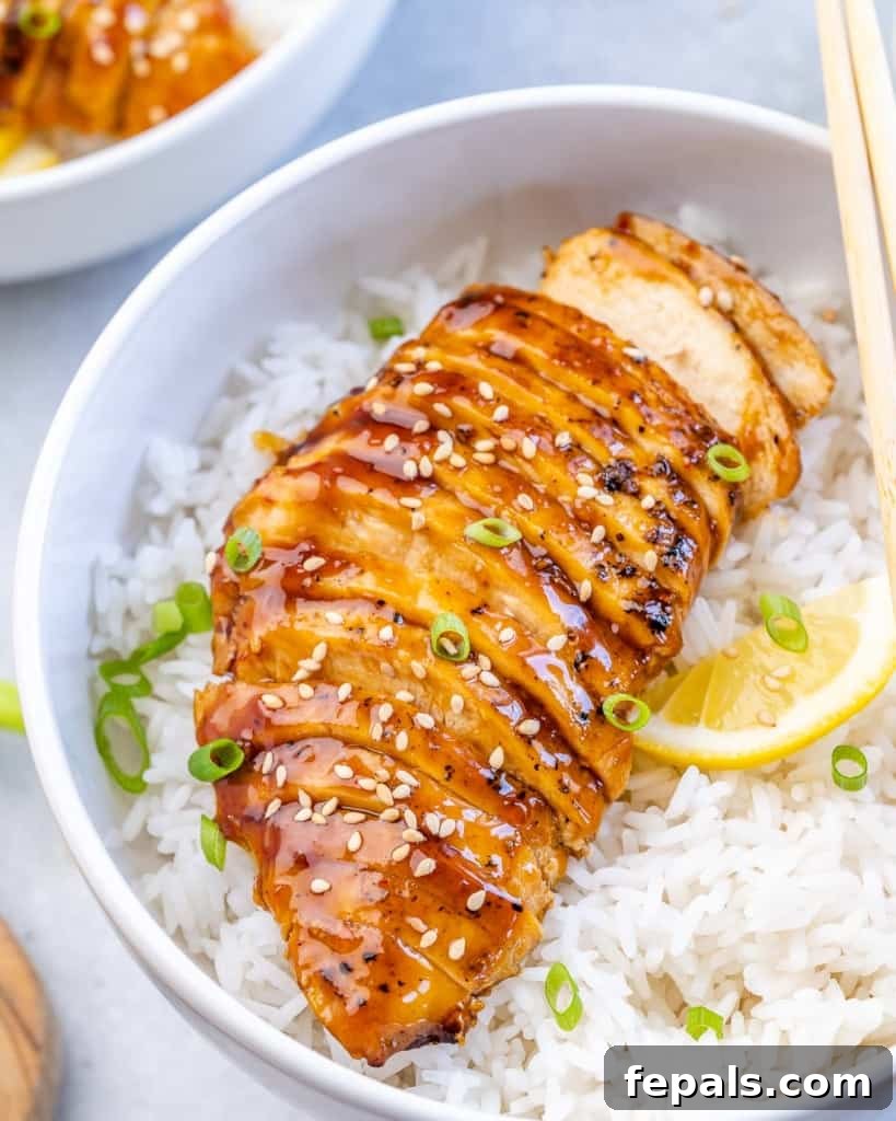 Side shot of perfectly sliced baked teriyaki chicken breast, glistening with sauce, served over a bed of fluffy white rice.