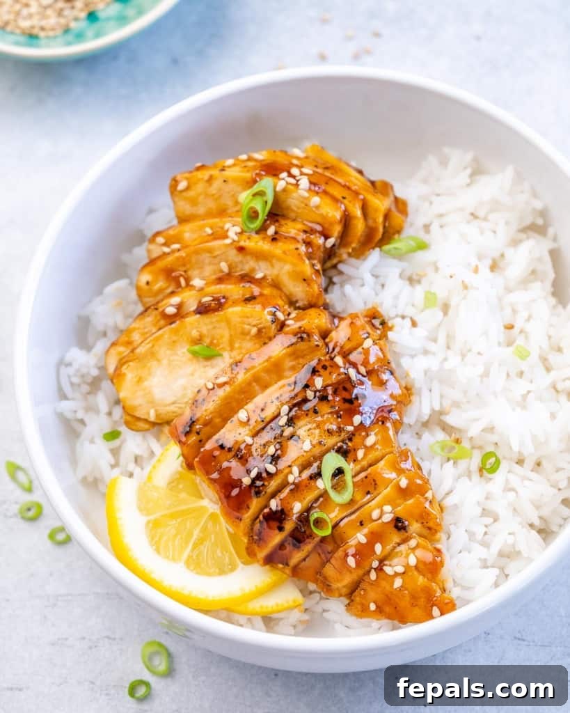 Side shot of perfectly sliced teriyaki chicken over white rice, garnished with fresh lemon wedges and green onions.