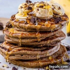 close up image of stacks of banana oatmeal pancakes topped with banana, shaved chocolate, and maple syrup drizzle