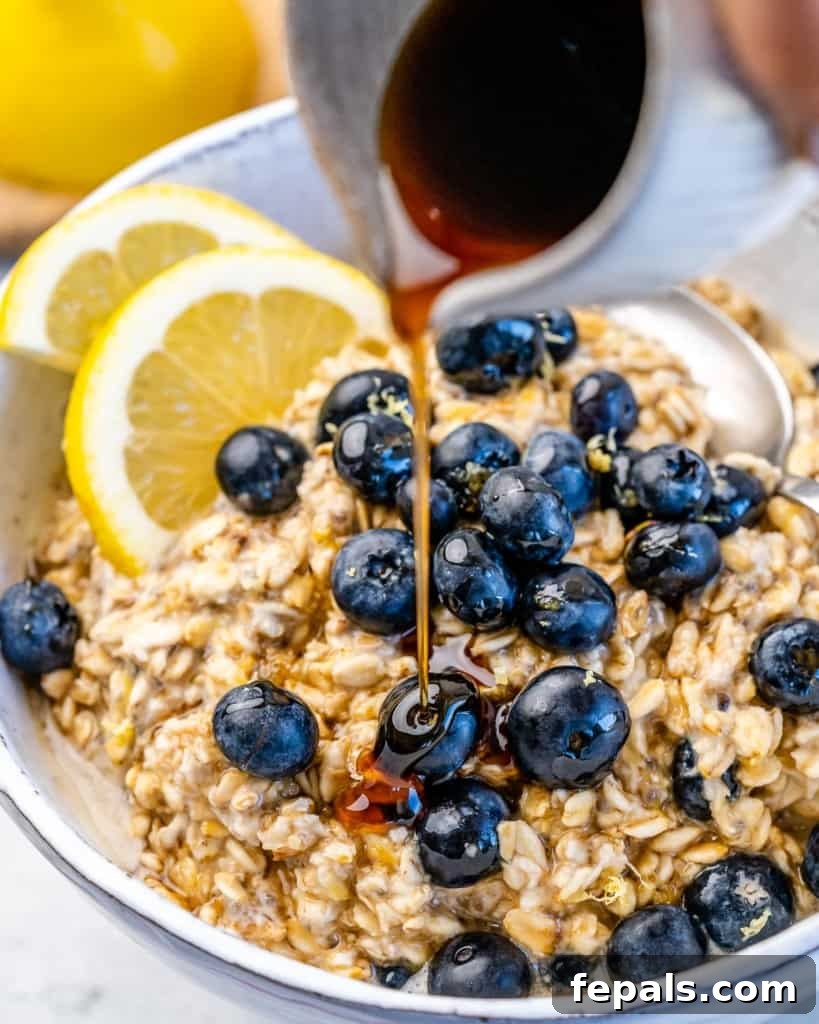 Berry Bliss Overnight Oats 9 Maple syrup being drizzled over a bowl of creamy blueberry overnight oatmeal, topped with fresh blueberries, enhancing its appeal.