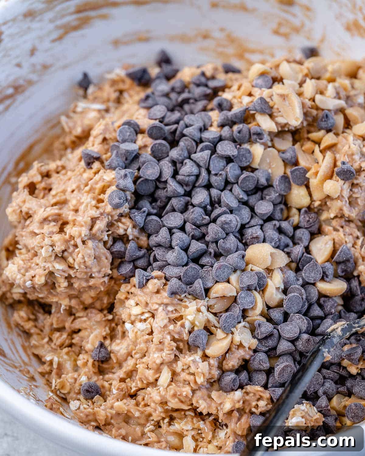 Wholesome Oat Energy Bars 4 A spoon mixing dark chocolate chips into a thick, textured oat mixture with peanut butter and peanuts, showing the comprehensive ingredient integration.