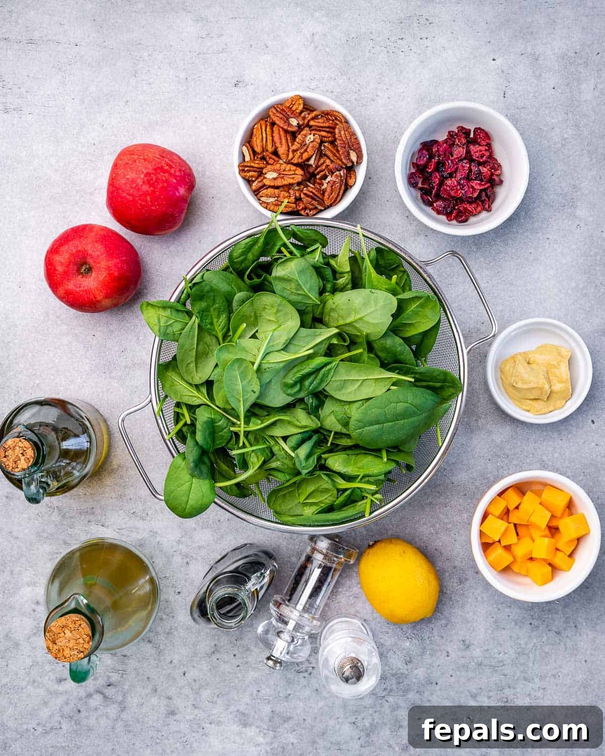A vibrant flat lay of all the fresh ingredients for Apple Pecan Salad: apples, cranberries, pecans, spinach, Dijon mustard, cheddar cheese, lemon, salt, pepper, maple syrup, and olive oil.