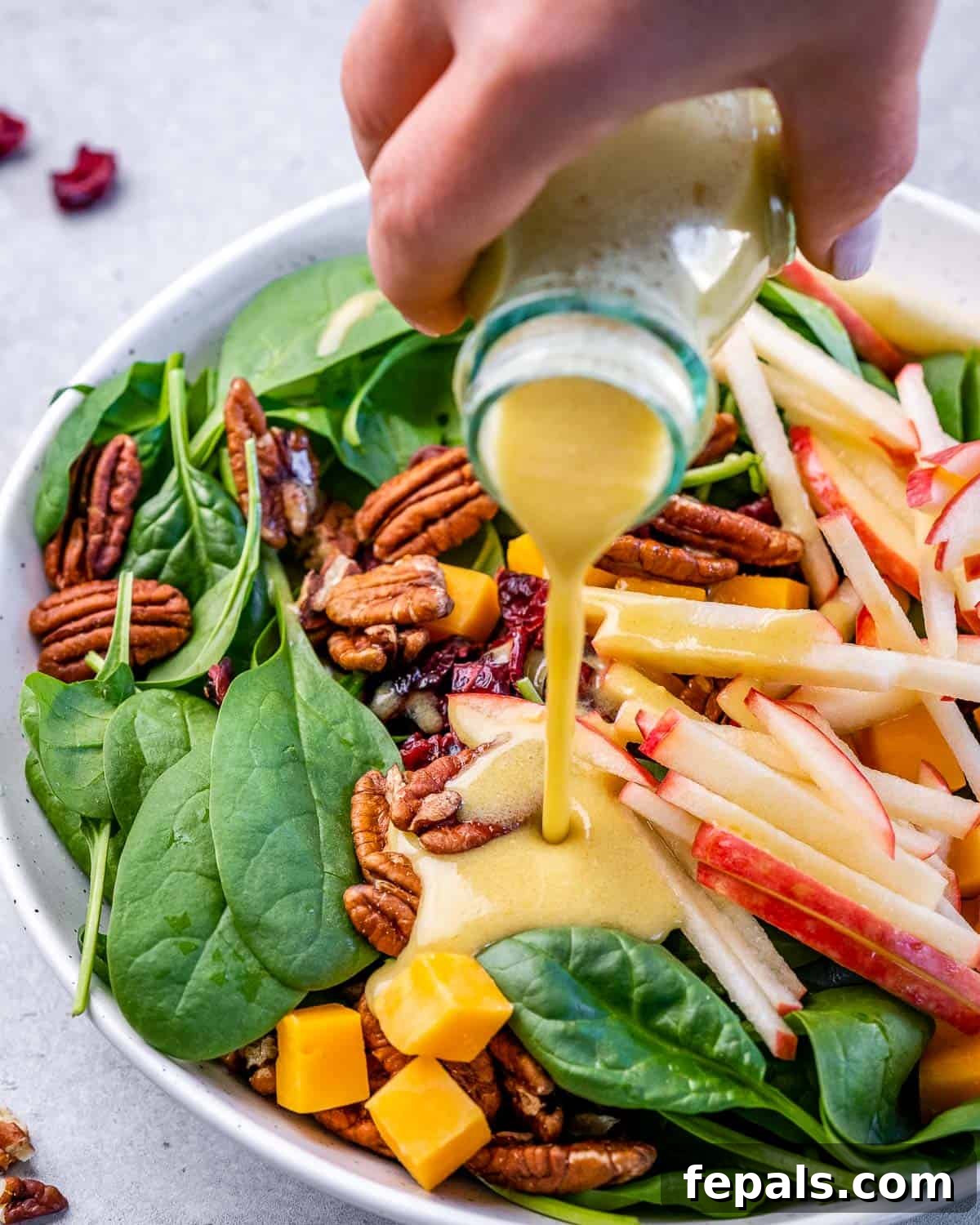 Close-up shot of maple vinaigrette being drizzled over a vibrant bowl of spinach salad with apples and pecans.