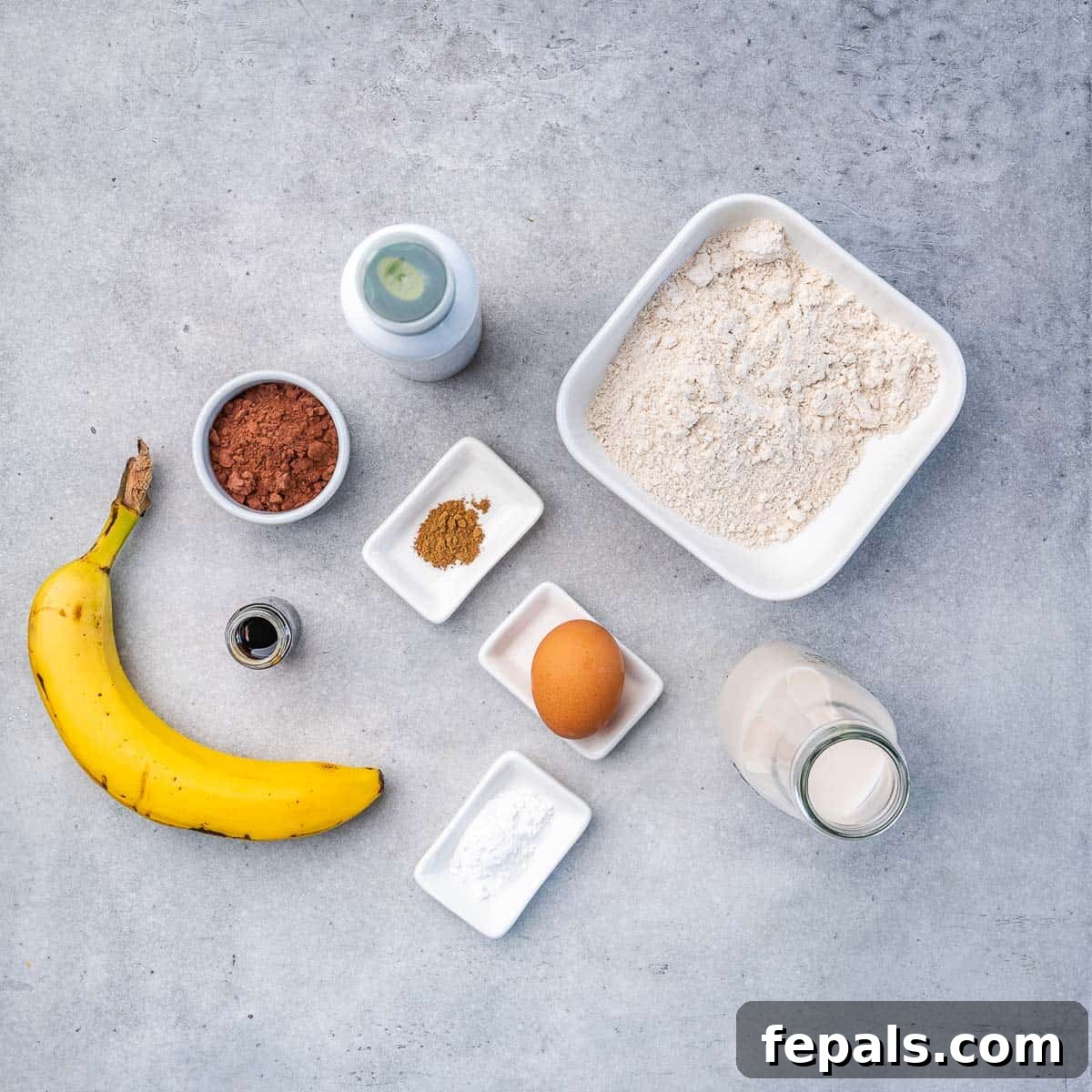 Chocolate Banana Pancakes A flat lay arrangement of ingredients for healthy chocolate banana pancakes: oat flour, cinnamon, cacao powder, a whole banana, an egg, milk, baking powder, and vanilla extract.