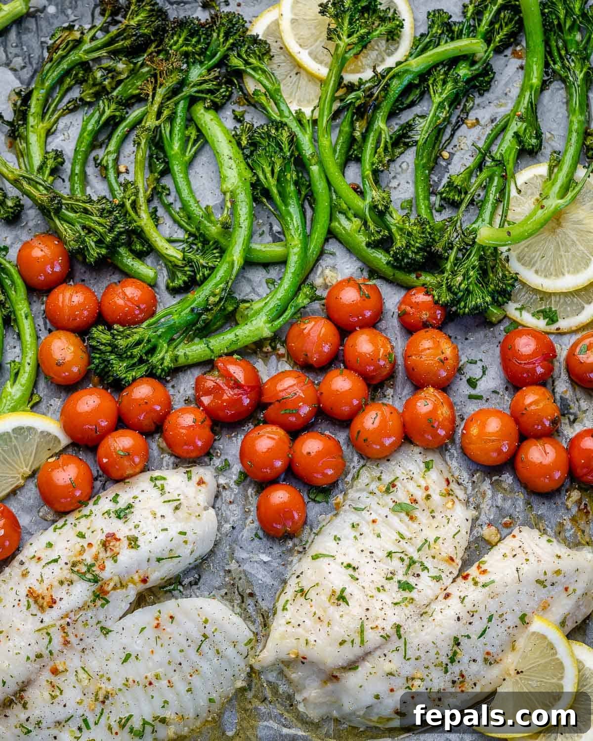 Baked Lemon Fish top view of tilapia fillets on the sheet pan with lemons, tomatoes, and broccolini