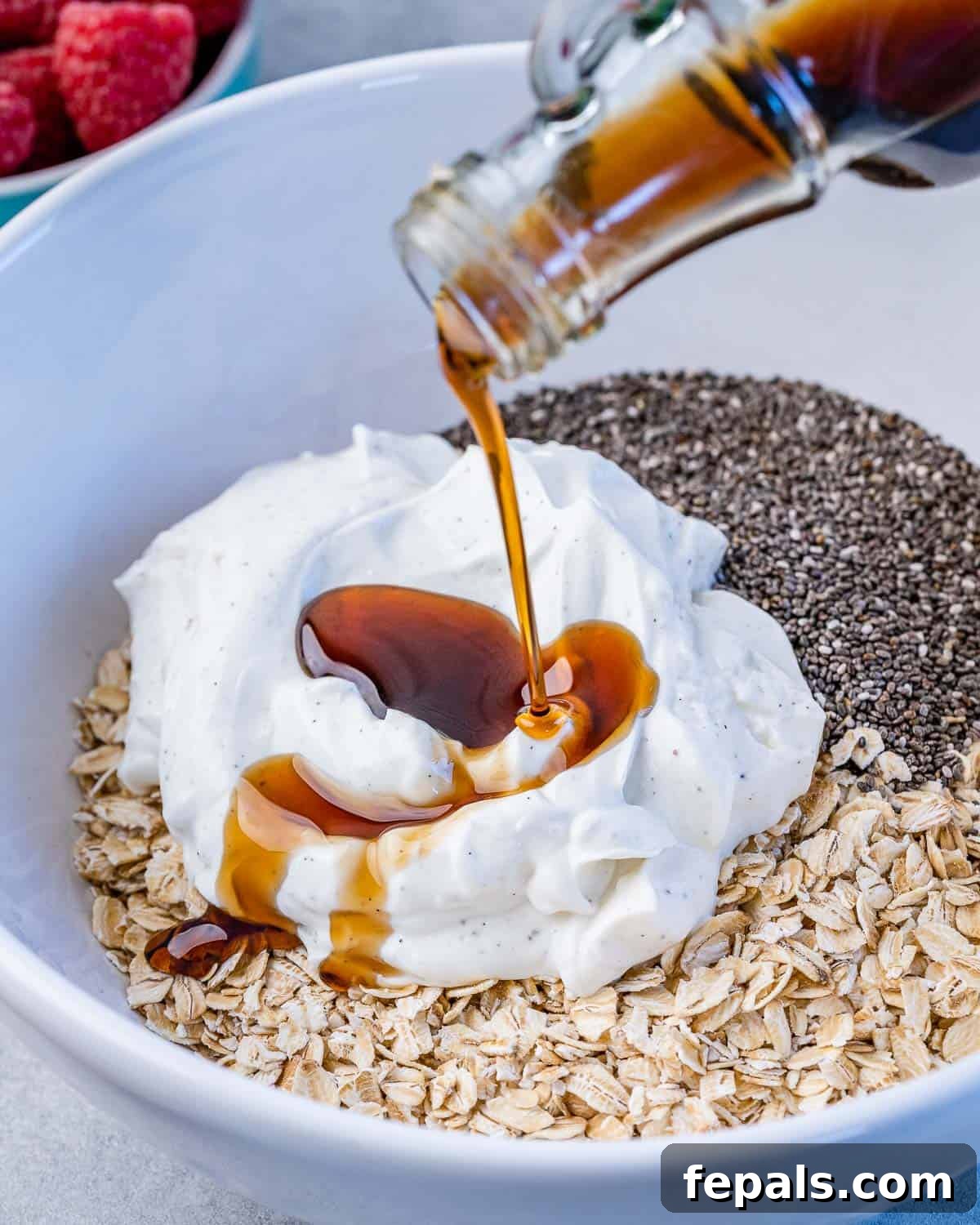 Dreamy PB&J Overnight Oats 5 maple syrup being poured over yogurt, oats, and chia seeds in a white bowl, the first step to making overnight oats